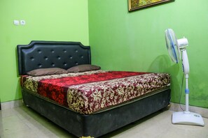 A bed with a dark headboard and a red and gold patterned bedspread. A white standing fan is next to the bed.