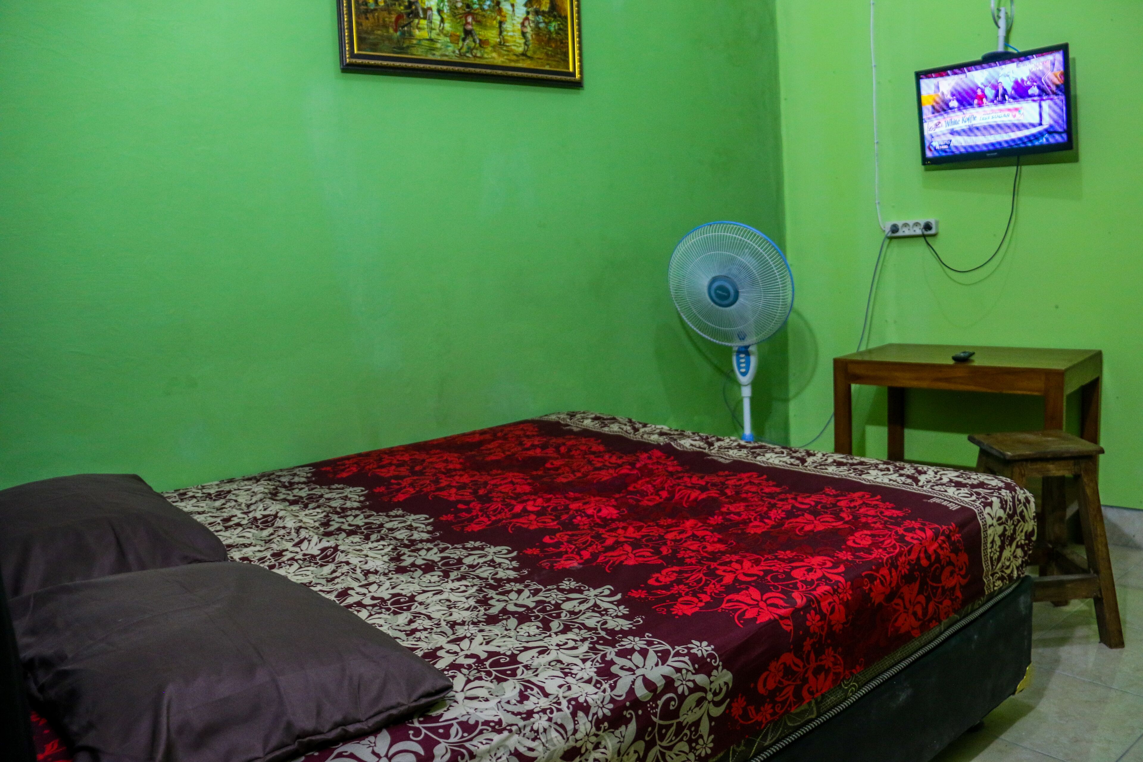 A room with a bed, a television mounted on the wall, a fan, and a small wooden table.