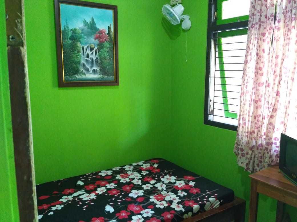 A room with a green wall, a bed with a floral bedspread, a painting of a waterfall, a fan, and a window with curtains.