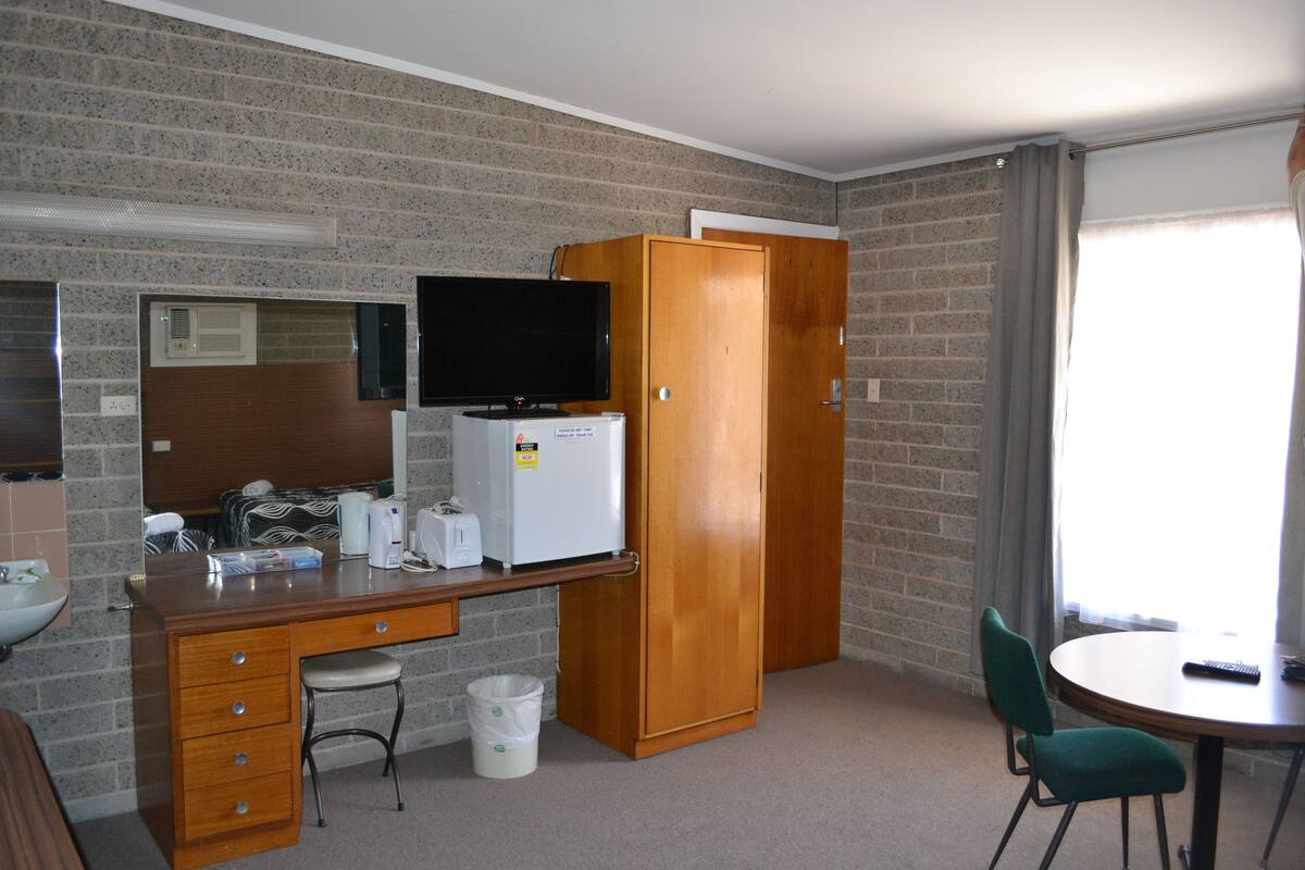A compact hotel room with a kitchenette, a single bed, a TV, a wooden wardrobe, and a small dining table with a chair.