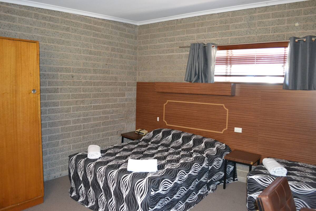 A room with a brick wall, a wooden door, a bed with a zebra-patterned bedspread, a small bedside table, and a window with curtains.