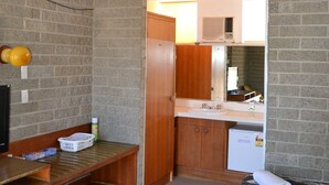 A compact hotel room with a kitchenette, a bed with a patterned bedspread, a wooden bench, and a trash bin.