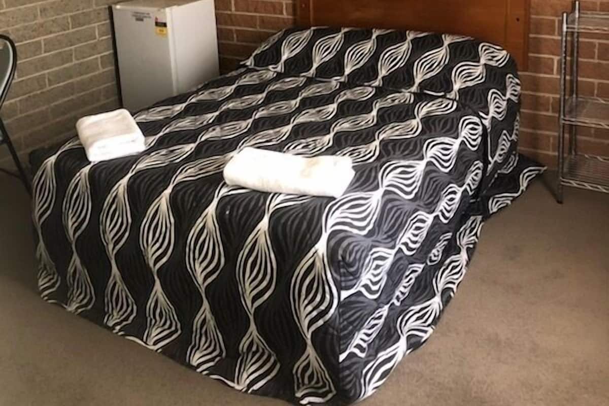 A small, single-bed room with a brick-patterned wall, a wooden headboard, a small air conditioning unit, and a metal shelf.