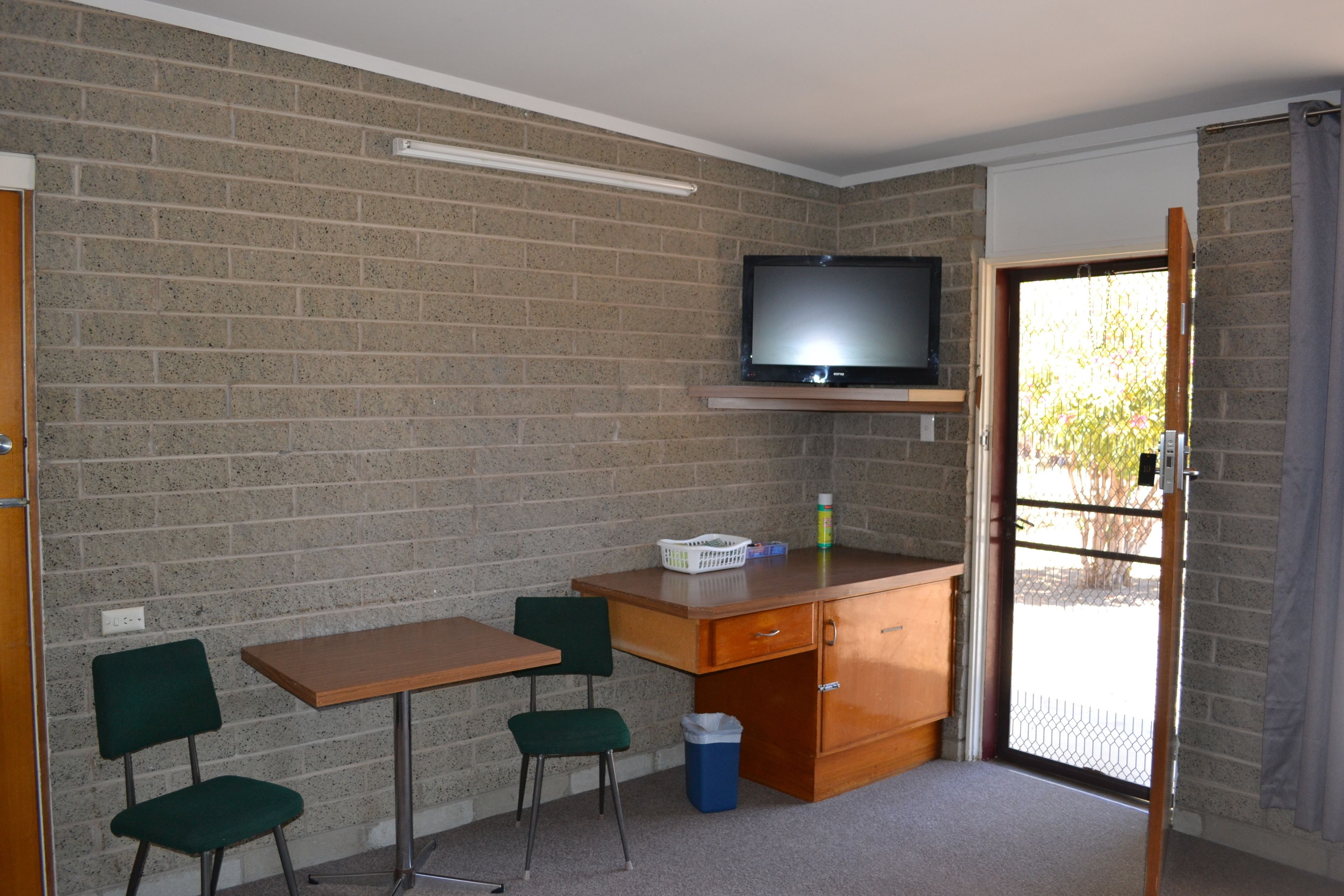 Una habitación con pared de ladrillo, un escritorio de madera, dos sillas verdes, un televisor sobre una estantería, un cubo de basura y una puerta que da al exterior.