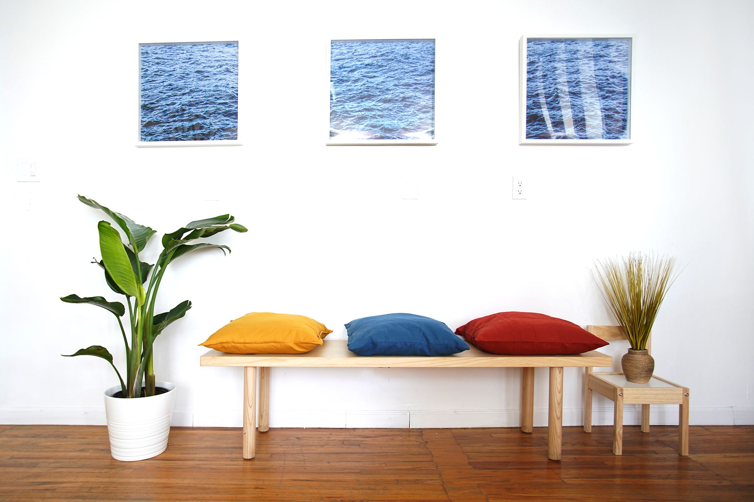 A room with a wooden bench, three cushions, a potted plant, and three framed pictures on the wall.