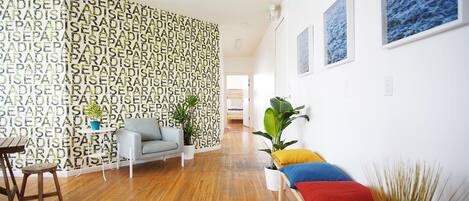 Uma sala de estar moderna com piso de madeira, um sofá cinza, uma mesa de centro de madeira e um banco colorido. Há plantas em vasos e quadros nas paredes.