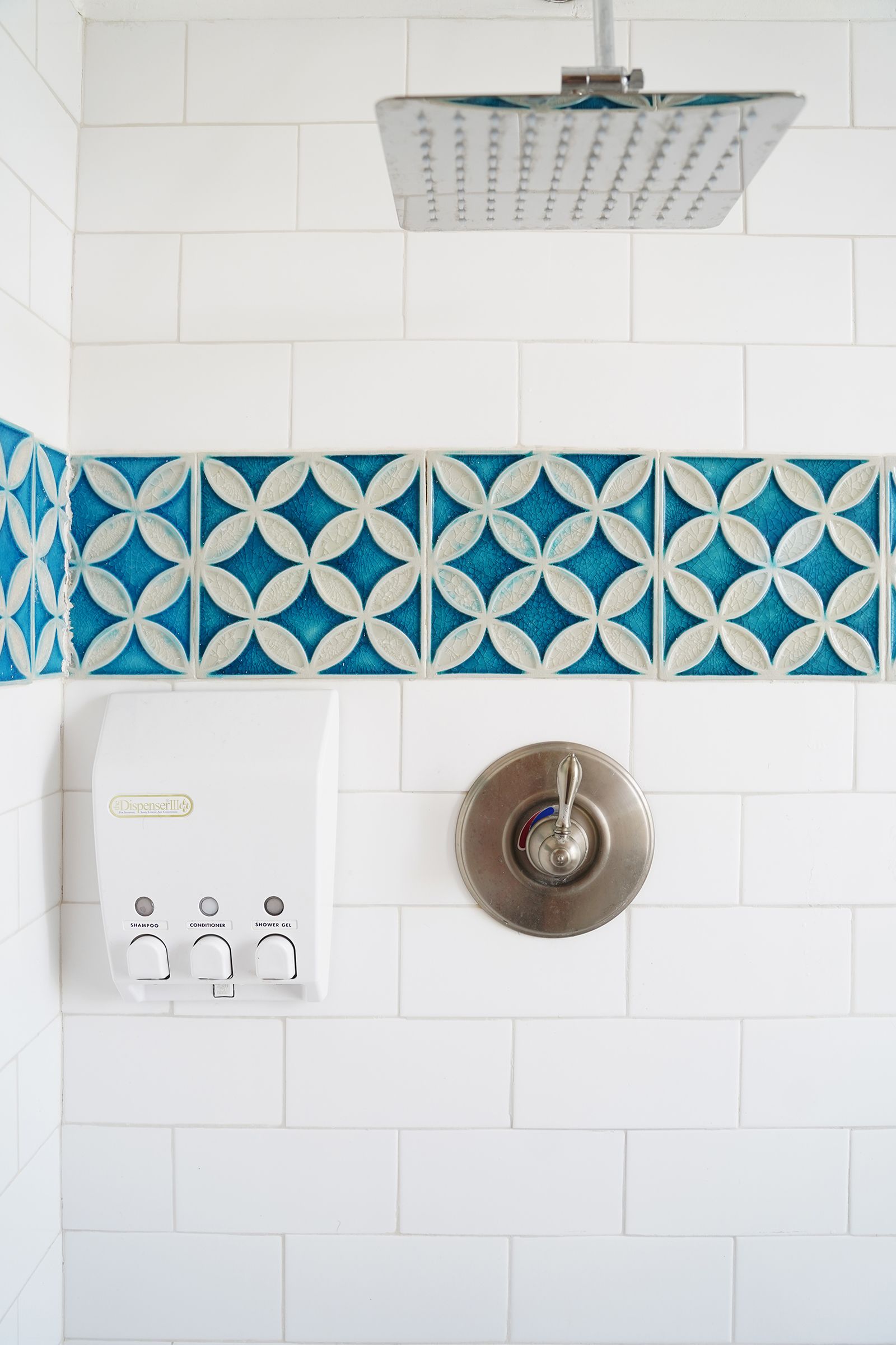 A bathroom with white tiles, a blue and white patterned shower curtain, and a dispenser with three buttons labeled "shampoo," "conditioner," and "shower gel."