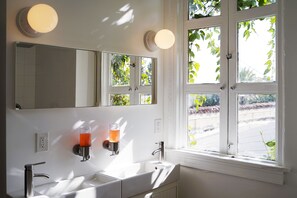 A bathroom with a double sink, a mirror, and a window with a view of greenery. - The Steady (Los Angeles)