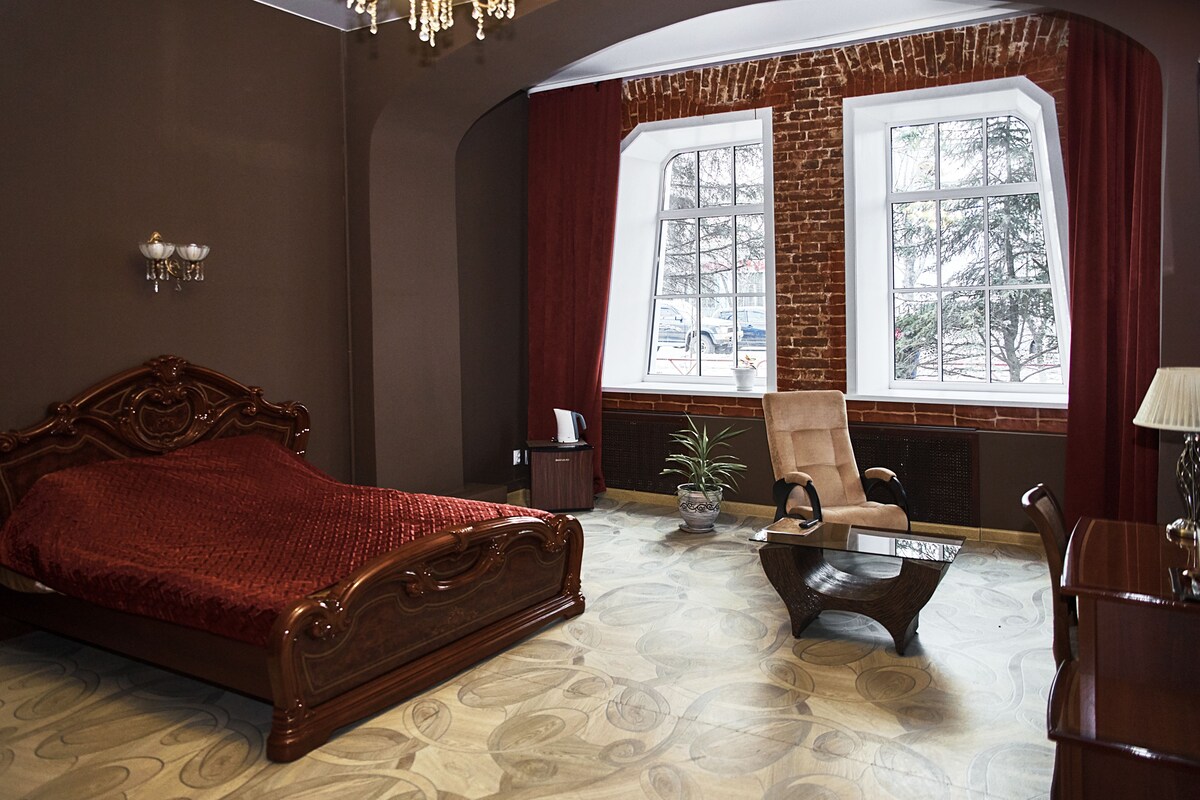 A bedroom with a large bed, a chair, a table, and a window with a view of trees.