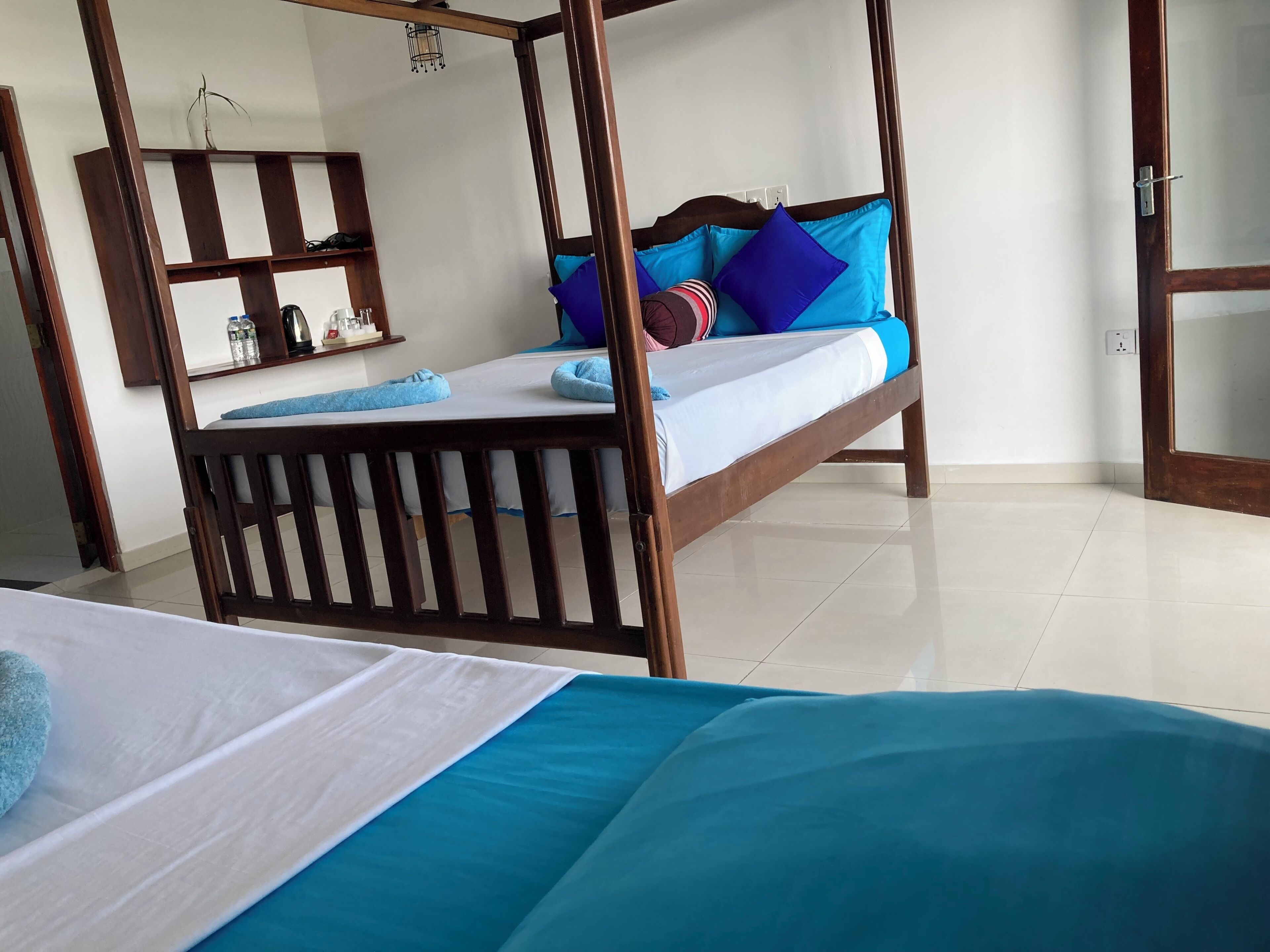 A room with a wooden four-poster bed, a small bedside table with a lamp, a mirror, and a shelf with toiletries.