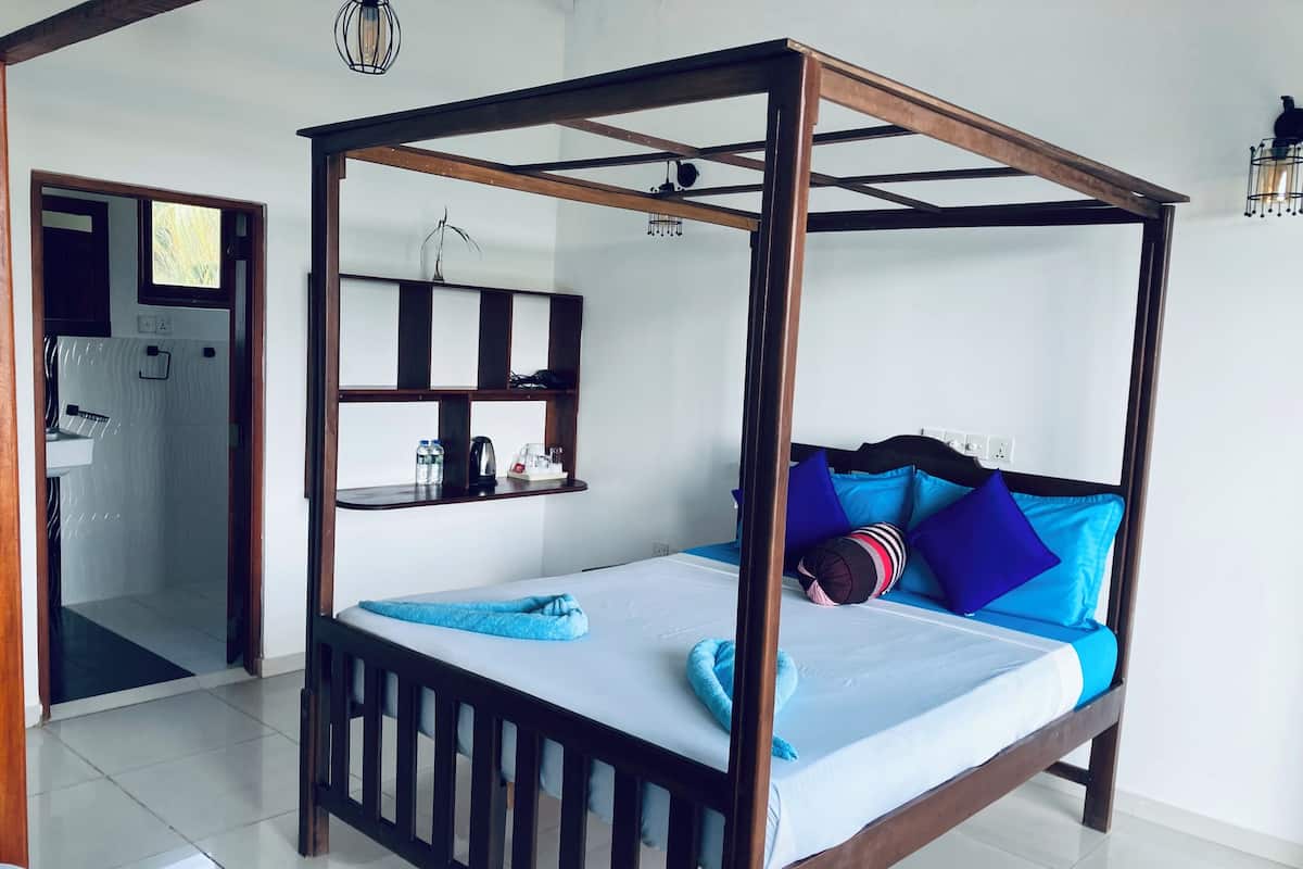 A four-poster bed with a wooden frame, a white mattress, and blue bedding. There is a shelf above the bed with a kettle, cups, and bottles. A small window is visible in the background.