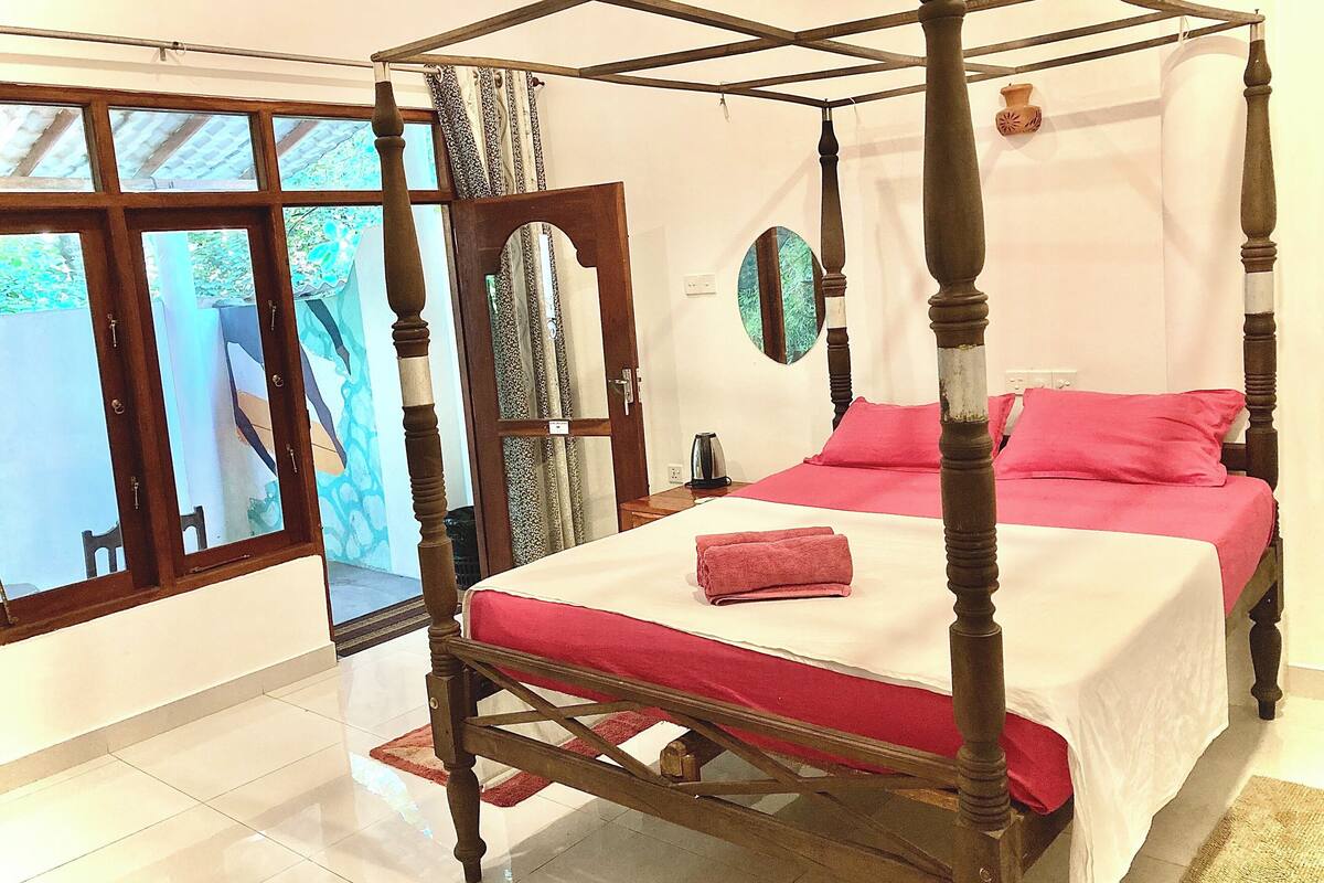 A four-poster bed with a red bedspread and pillows, a wooden floor, a window with a view of greenery, and a wall-mounted mirror.