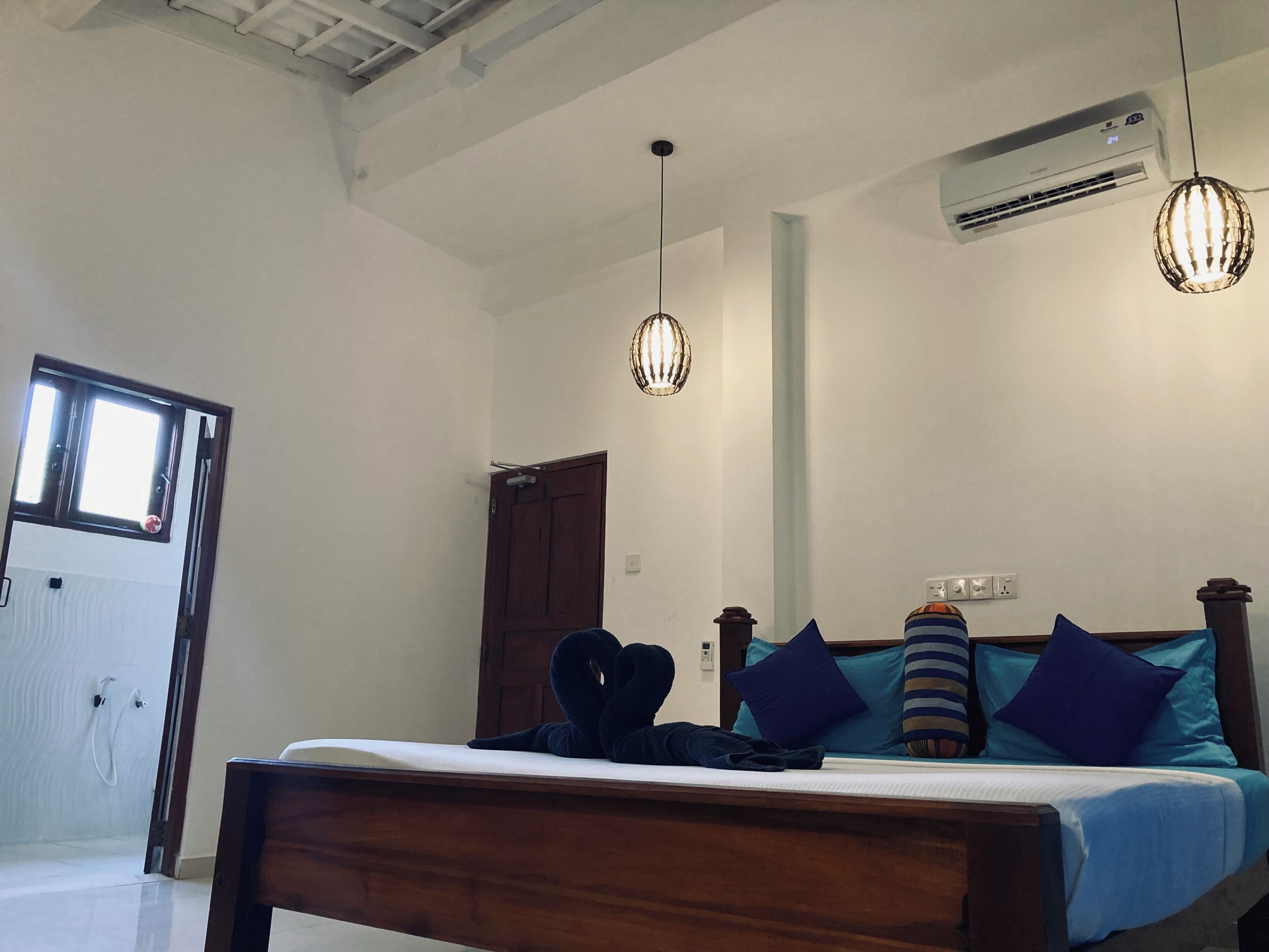 A bedroom with a bed, two hanging lights, an air conditioning unit, a door, and a window.