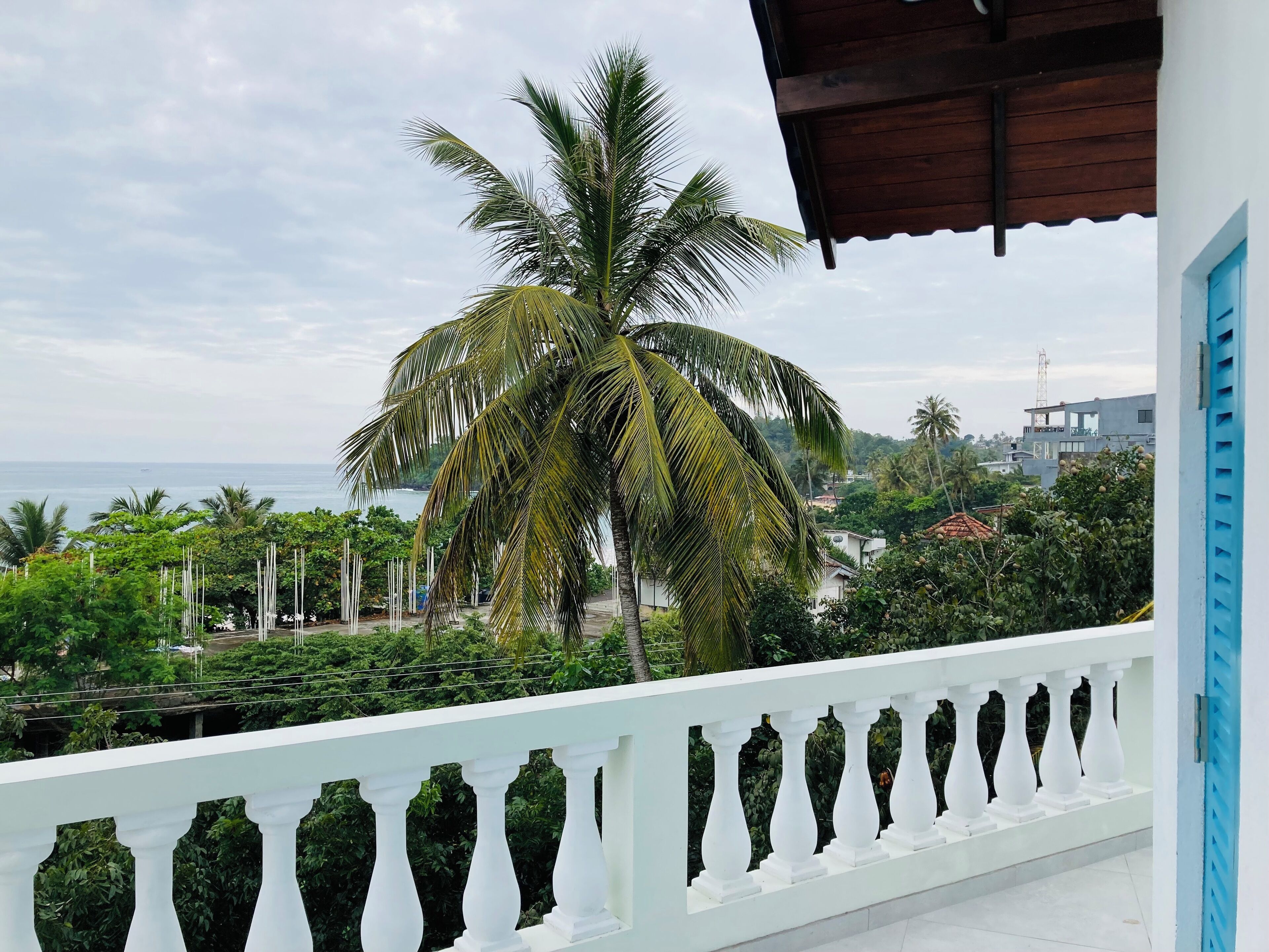 Un balcon doté de balustrades blanches, donnant sur une zone côtière bordée de palmiers et offrant une vue sur la mer.