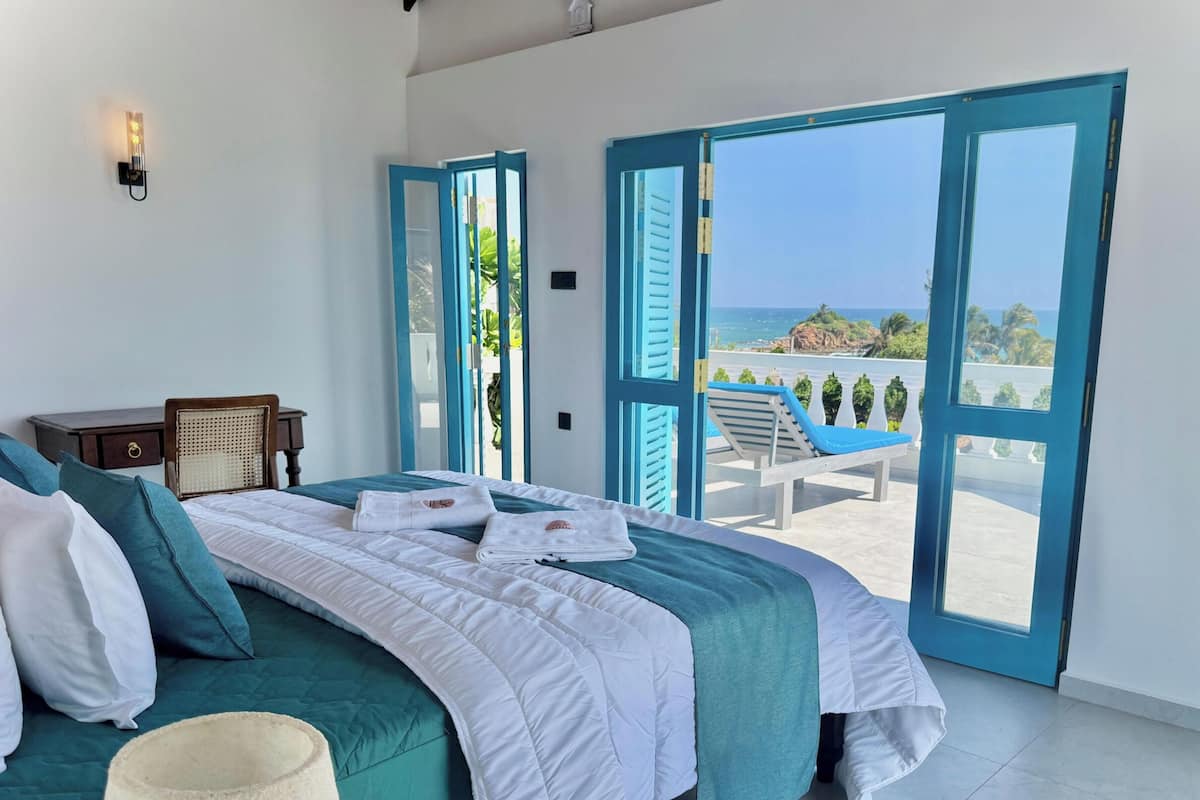 Un dormitorio con cama, mesita de noche, ventana con persianas azules y vista al océano.
