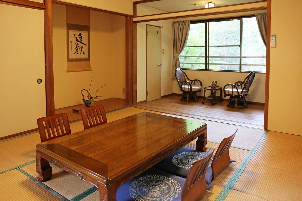 A traditional Japanese-style room with tatami flooring, a wooden dining table, and a view of a garden through a large window.
