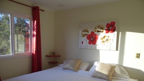 A bedroom with a large bed, a window with red curtains, a wall-mounted artwork of flowers, and a small shelf with a plant. - Villanita (Punta Arenas)
