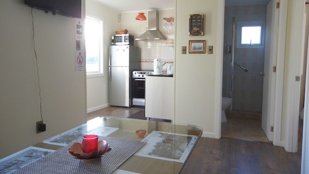 A compact kitchen with a refrigerator, microwave, stove, and sink. There is a dining area with a glass table and a red candle. - Villanita (Punta Arenas)