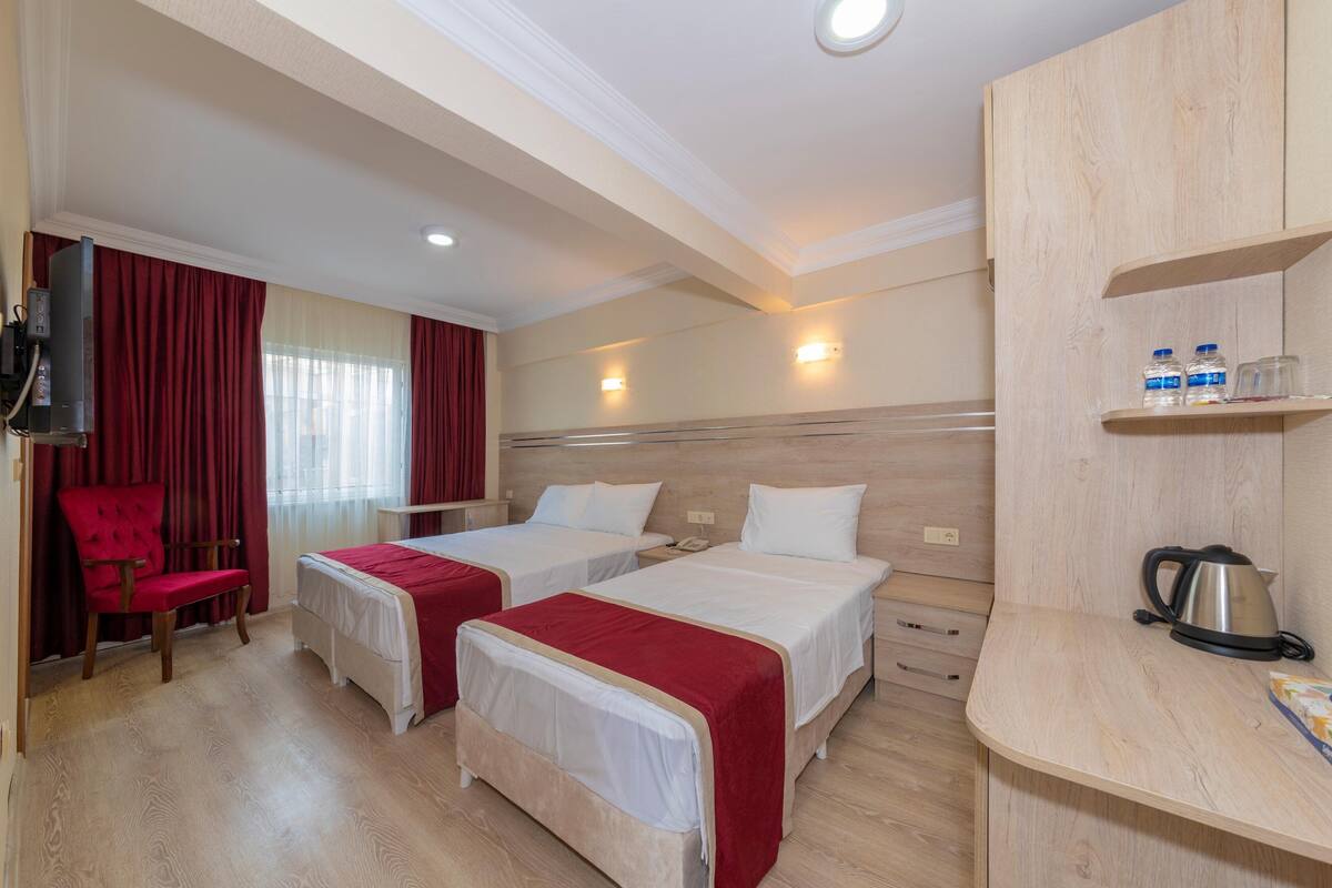 A hotel room with two beds, a red chair, a wooden desk, and a kettle.