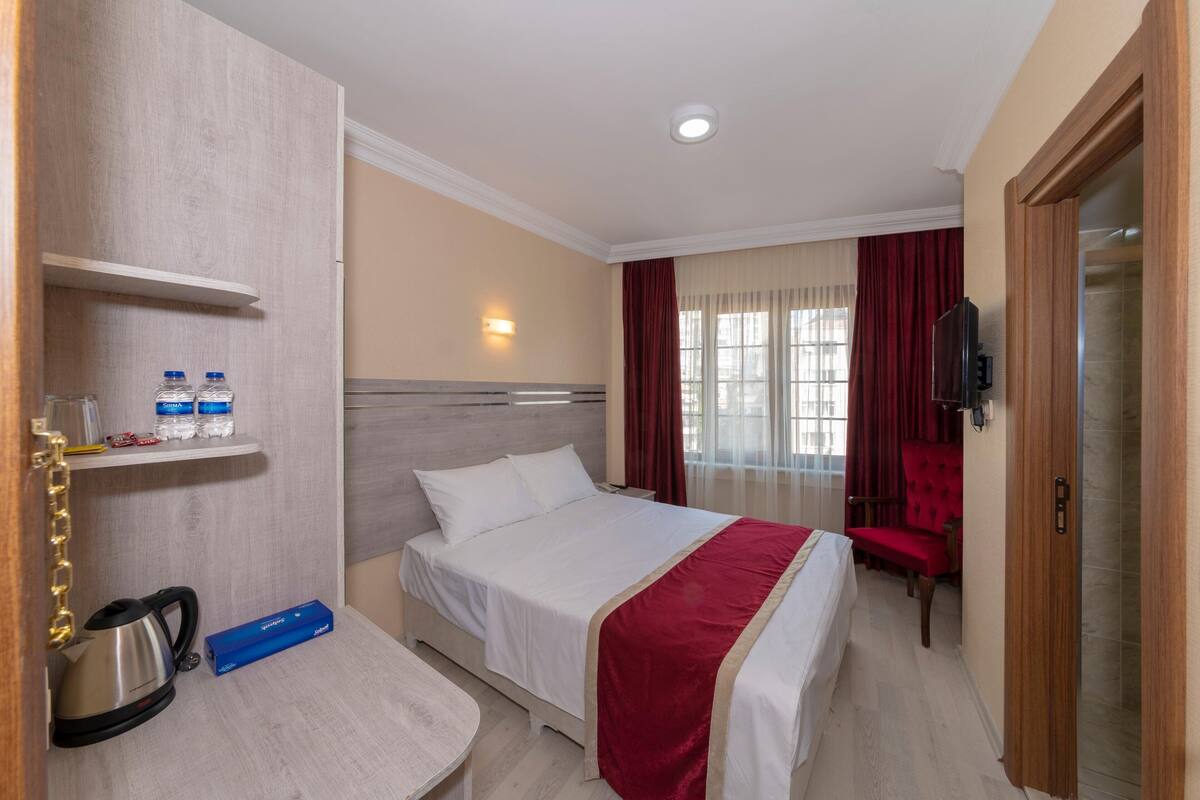 A hotel room with a bed, a desk with a kettle, a shelf with toiletries, and a TV.