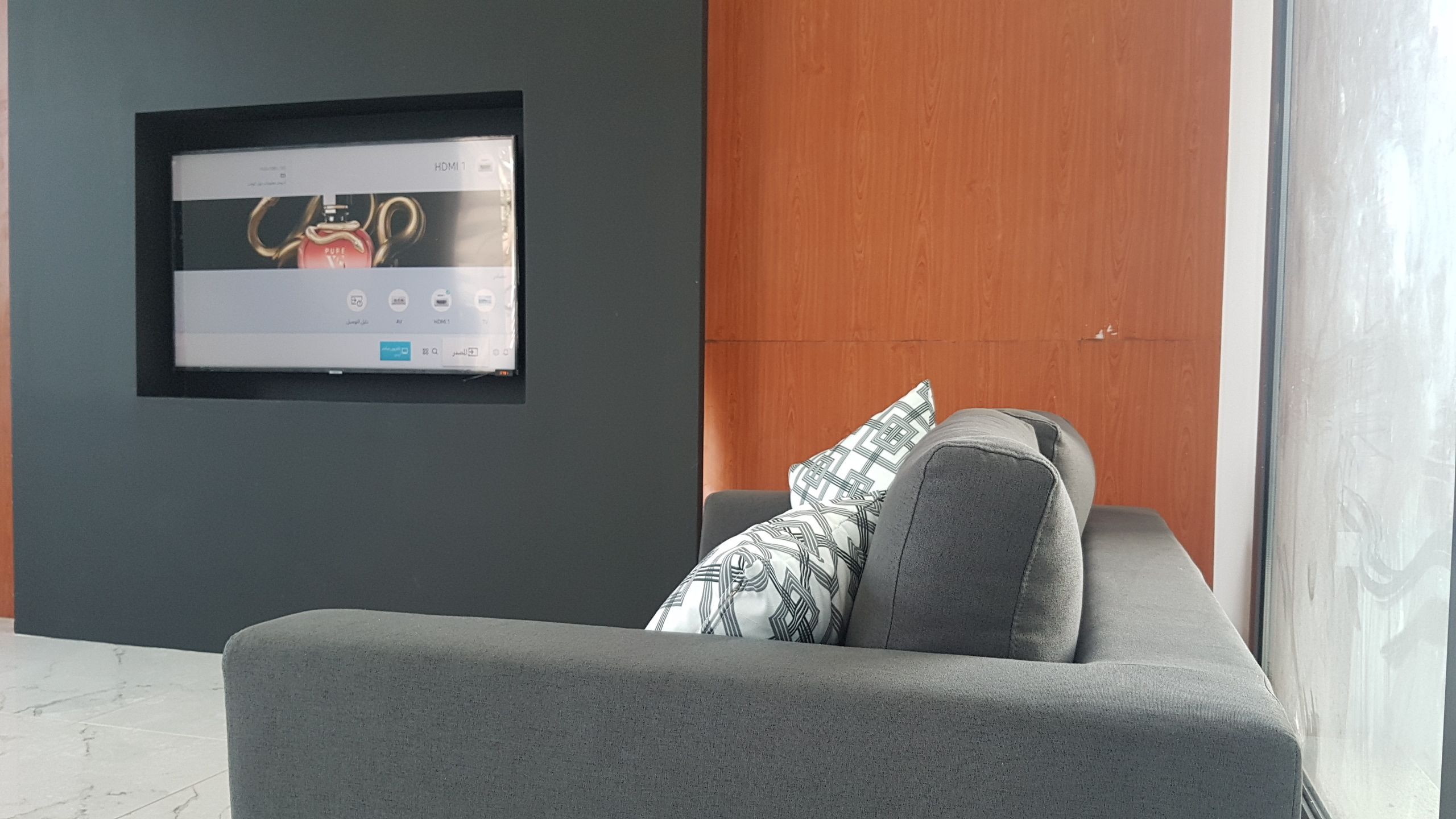 Een moderne woonkamer met een grijze bank, een wandgemonteerde televisie en een houten paneelwand.