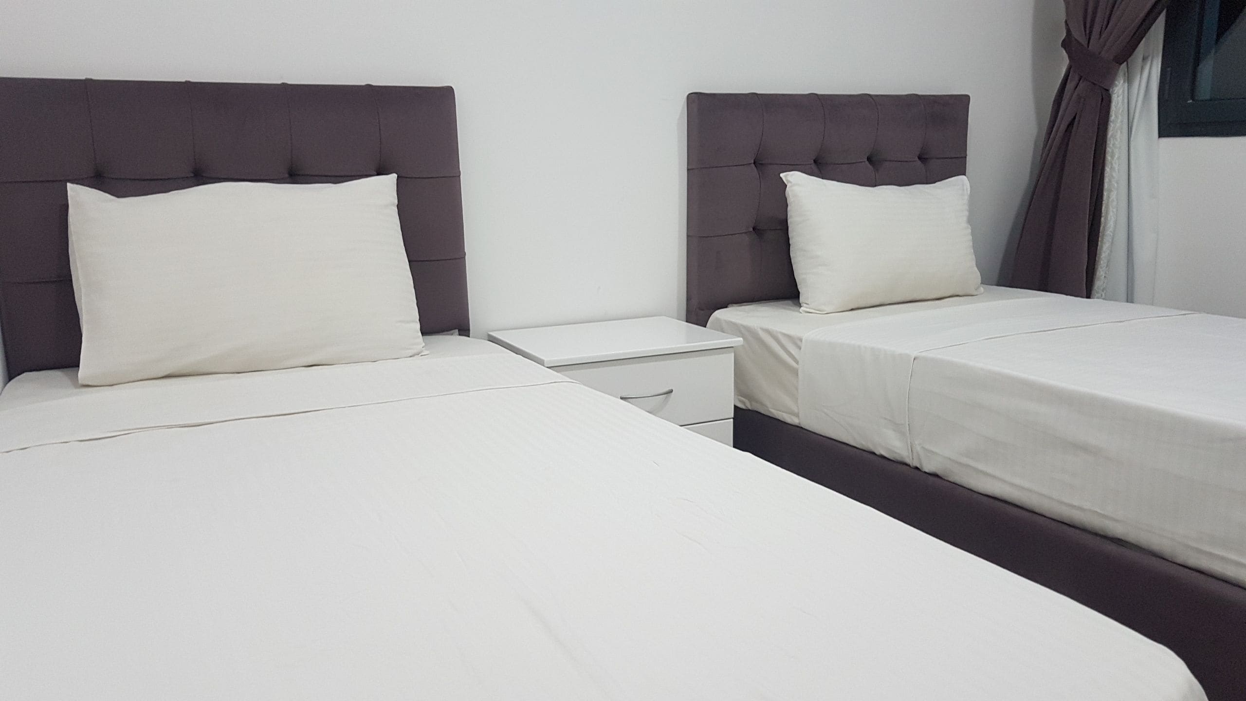 A hotel room with two beds, each with a headboard and a pillow. There is a nightstand between the beds.