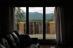 A room with a black leather sofa facing a large window with a view of mountains and a wooden fence.
