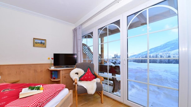 A hotel room with a bed, a chair, a TV, and a large window with a snowy view.