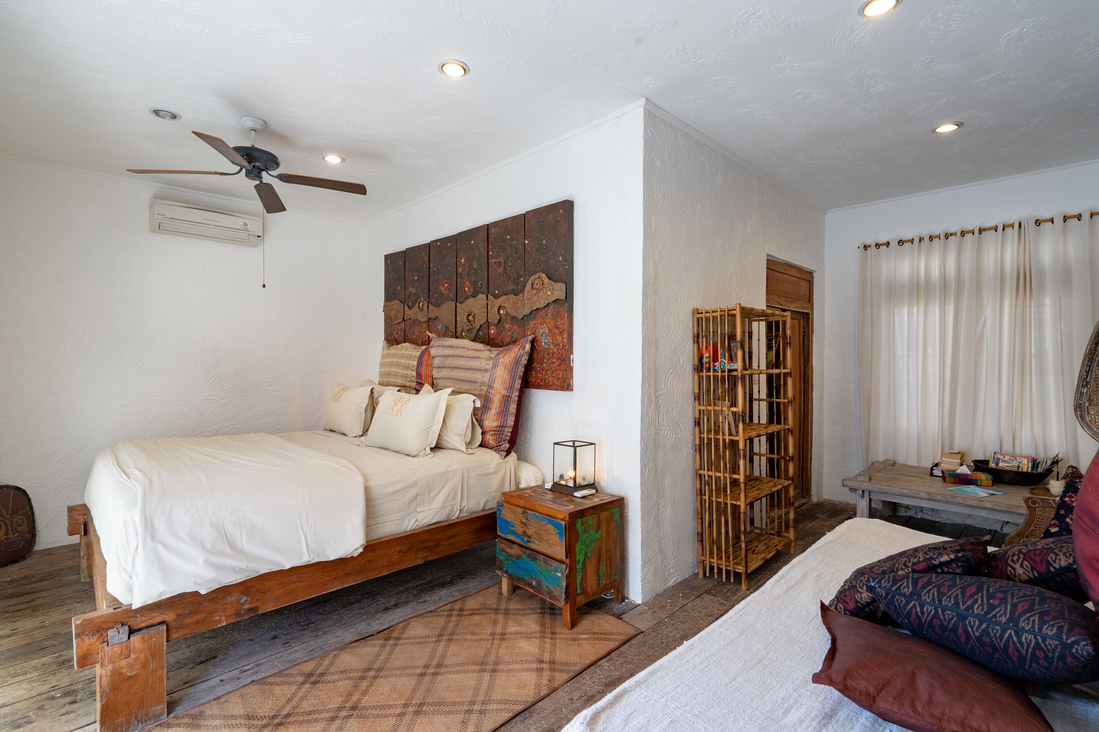 Un dormitorio con cama, ventilador de techo, aire acondicionado de pared, estantería de madera y una pequeña mesa con lámpara.