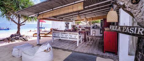 A beachside villa with a wooden deck, outdoor seating, and a sign reading "VILLA PASIR".
