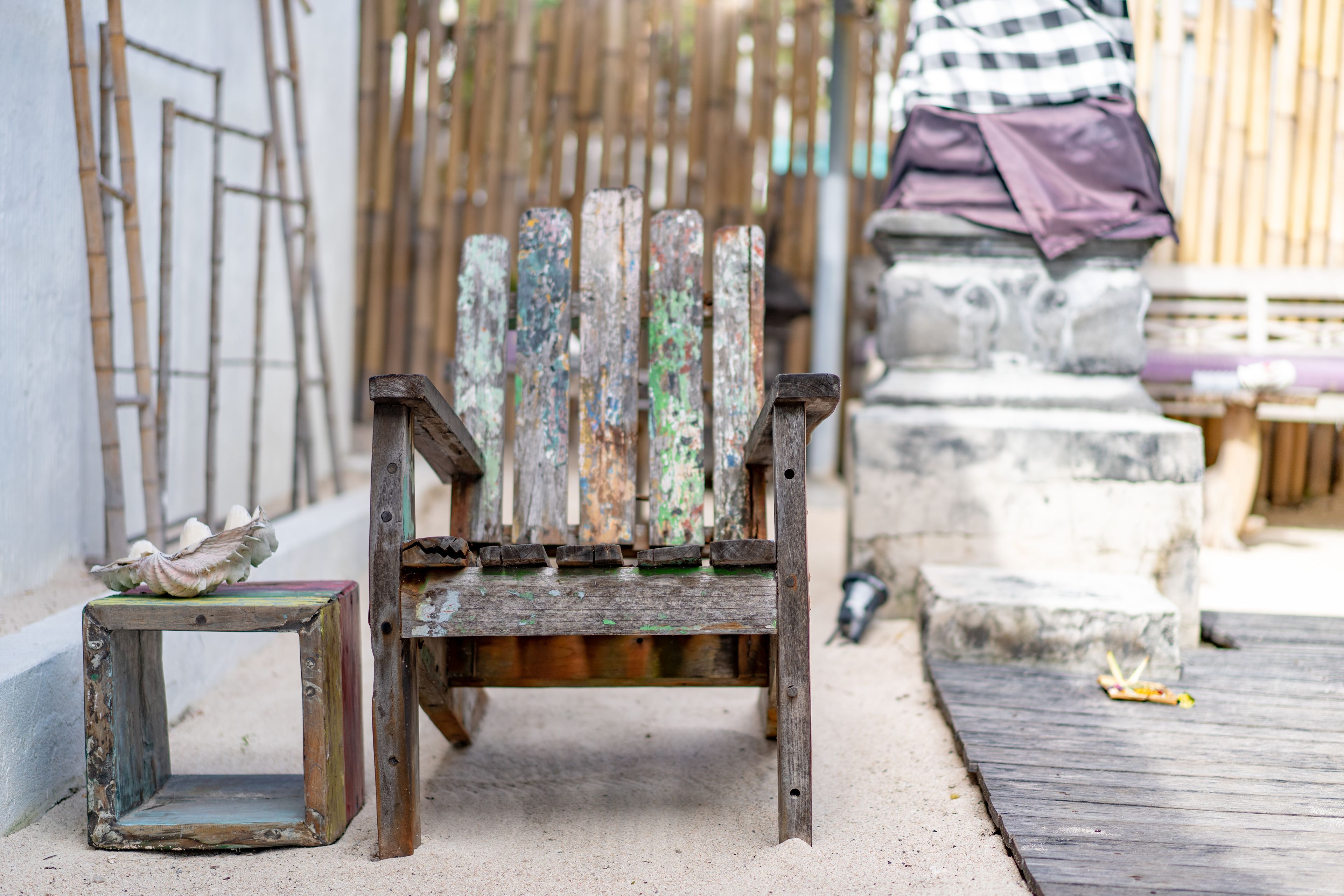 A rustic wooden chair with peeling paint, a small square stool, and a concrete bench in a sandy outdoor area.