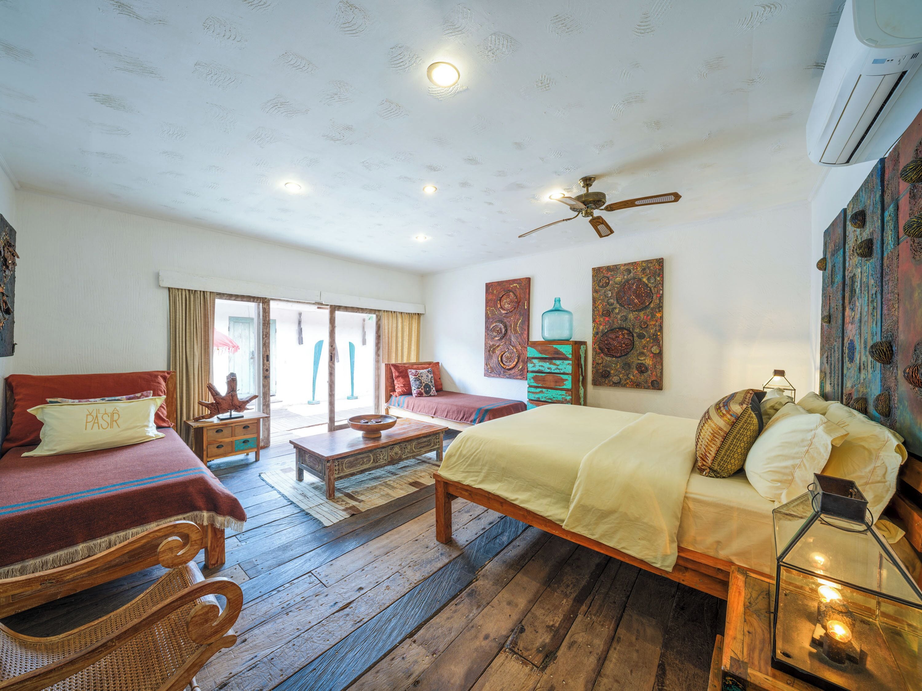 A room with a bed, two sofas, a wooden coffee table, a ceiling fan, and artwork on the walls.