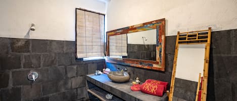 Un baño con pared de piedra, un espejo grande, una escalera de madera y una ventana con persianas.