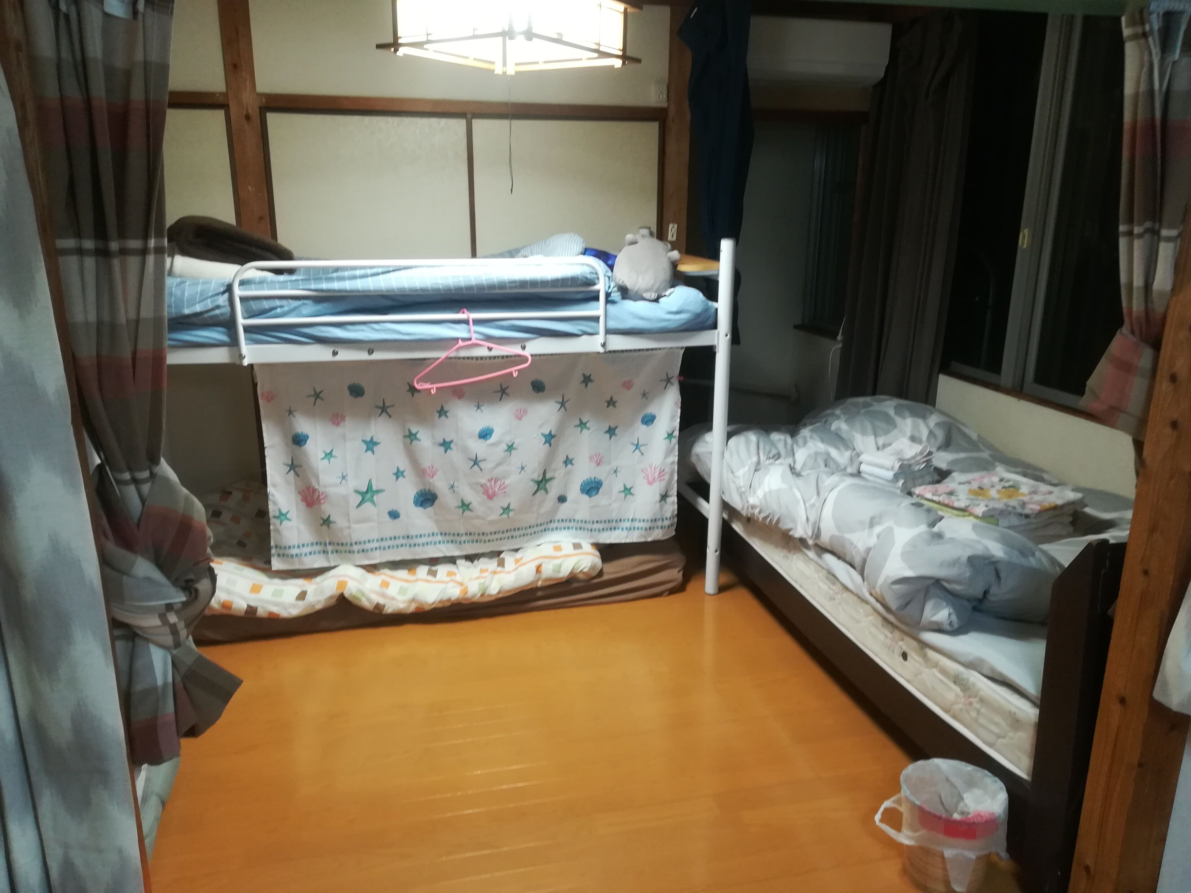 A dormitory room with bunk beds, a wooden floor, and a curtain-covered window.
