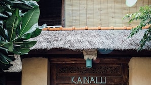 Una puerta de madera con el nombre "Kainali" grabado encima, ubicada en un edificio rústico con techo de paja.
