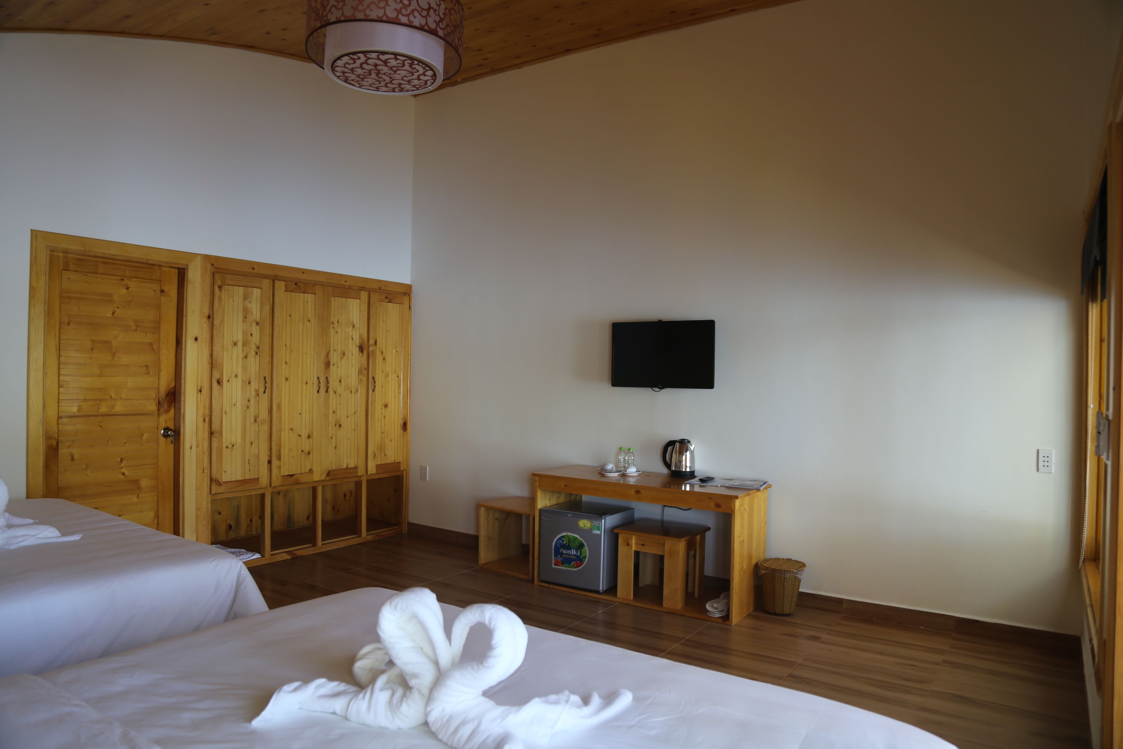 Habitación de hotel con dos camas, un armario de madera, un escritorio con televisor, un refrigerador y una pequeña mesa.