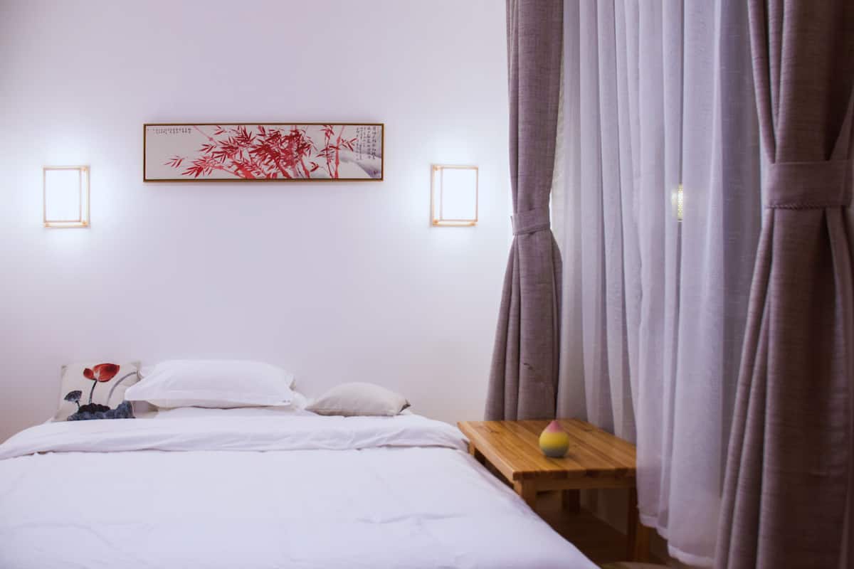 Una cama bien tendida con sábanas blancas, una pequeña mesita de noche con una lámpara y una obra enmarcada sobre la cama.