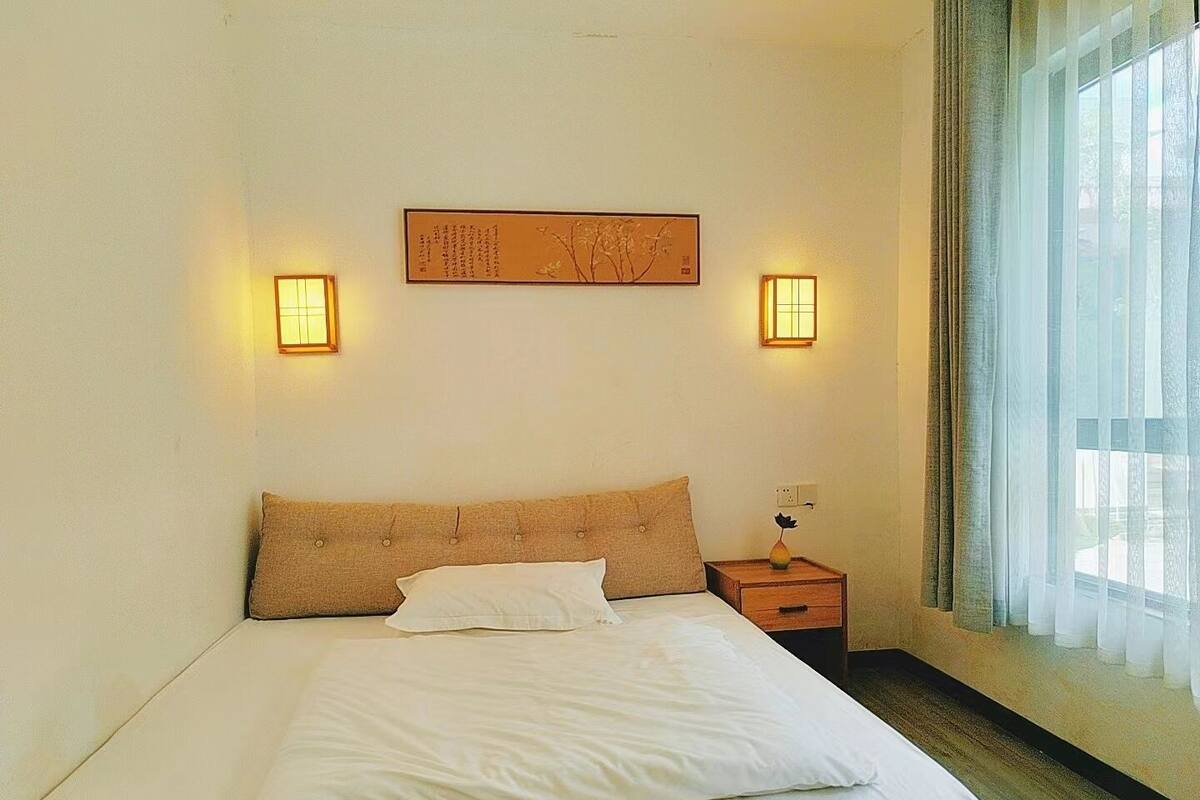 Una habitación de hotel con una cama, dos lámparas de pared, una mesita de noche, un florero y una ventana con cortinas traslúcidas.