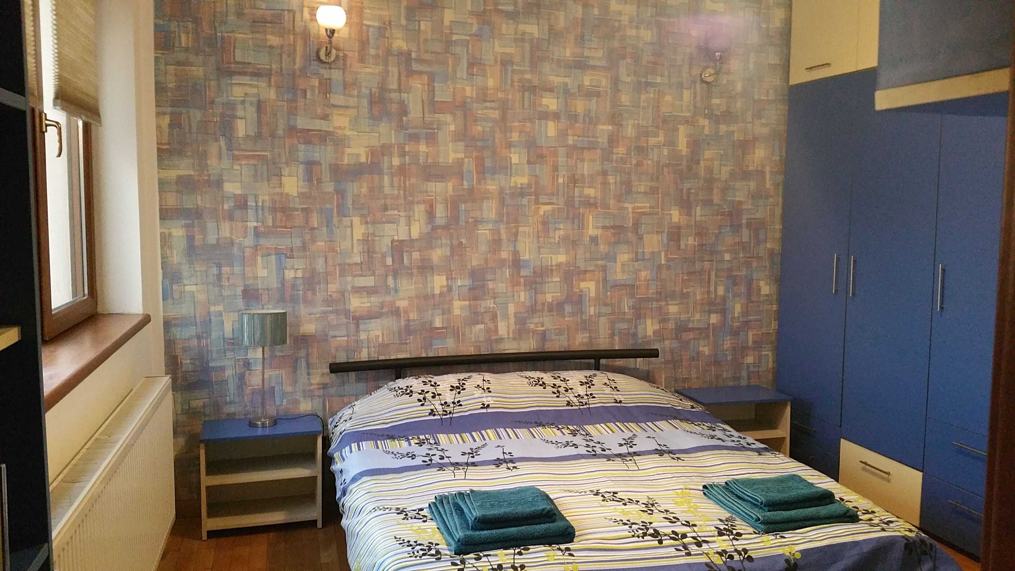 Un dormitorio con pared estampada, una cama con colcha a rayas, dos toallas verdes, una mesita de noche, una ventana y un armario.