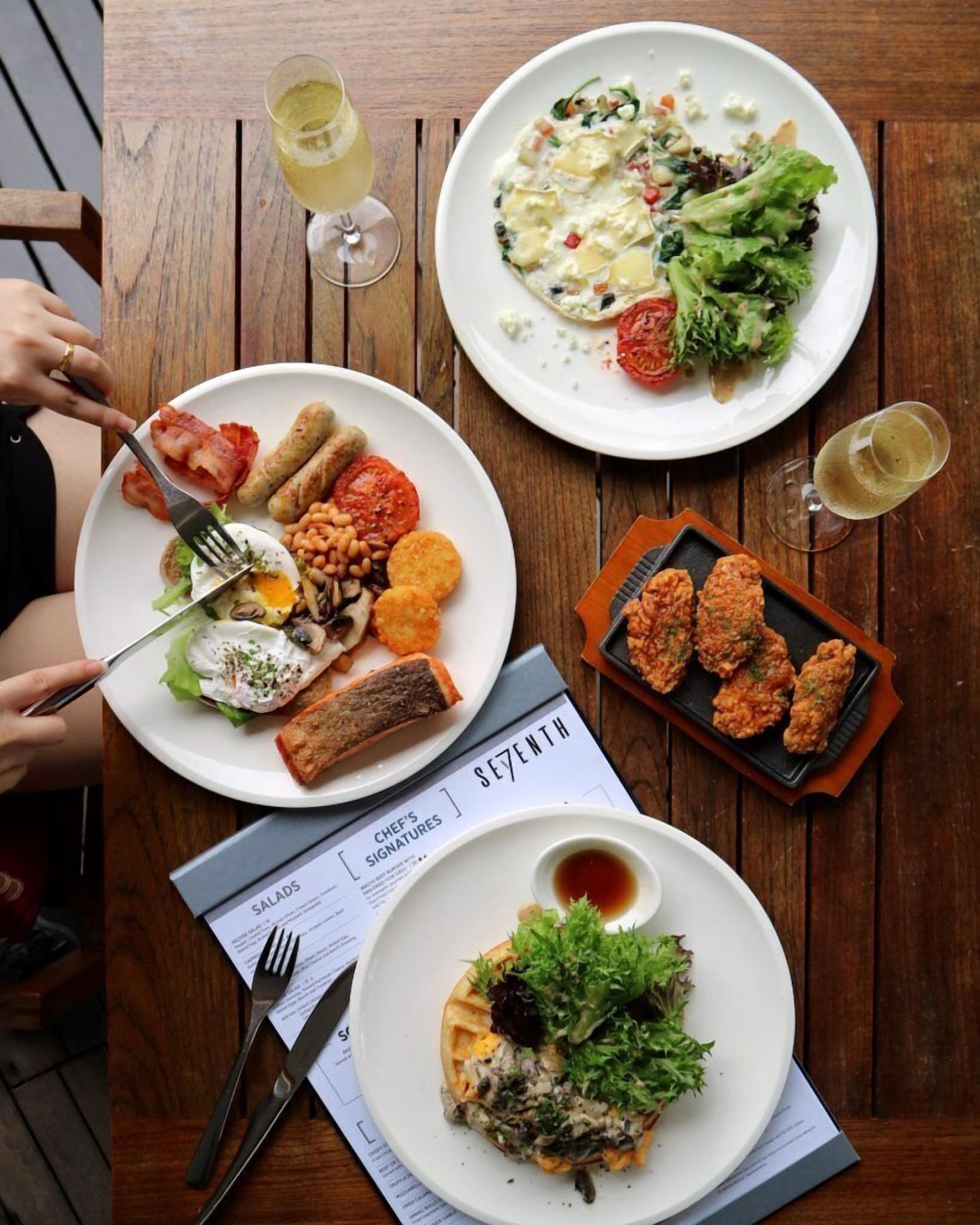 A wooden table with three plates of food, a menu, and two glasses of white wine.