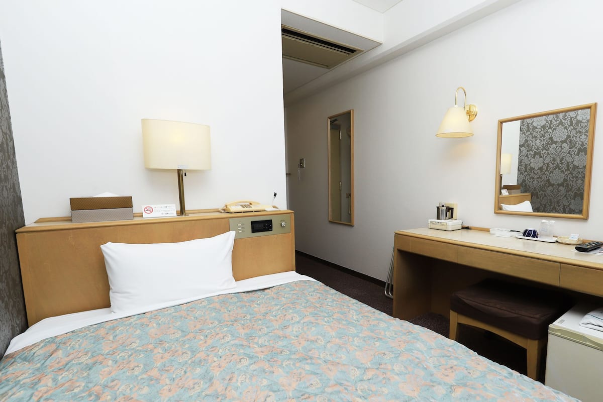 A hotel room with a bed, bedside table, lamp, mirror, and a desk with a chair.