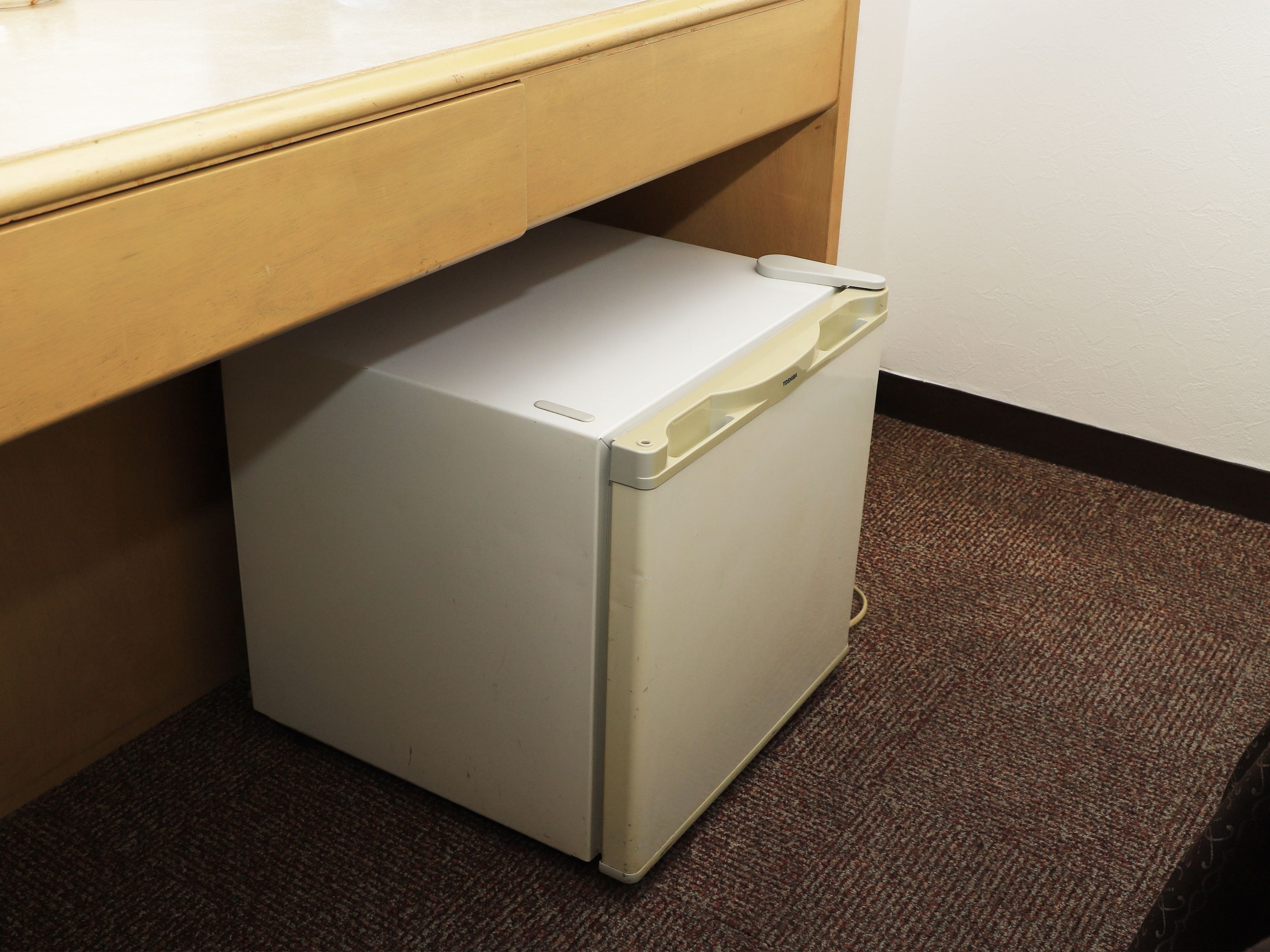 A compact, white refrigerator under a wooden counter in a room with a brown carpet.