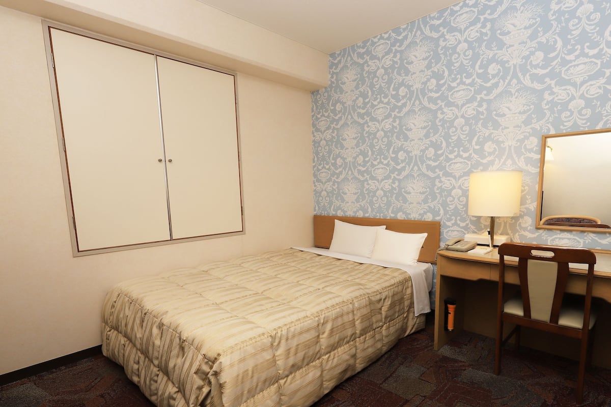 A hotel room with a bed, a desk with a lamp, a chair, a wardrobe, and a patterned wallpaper.