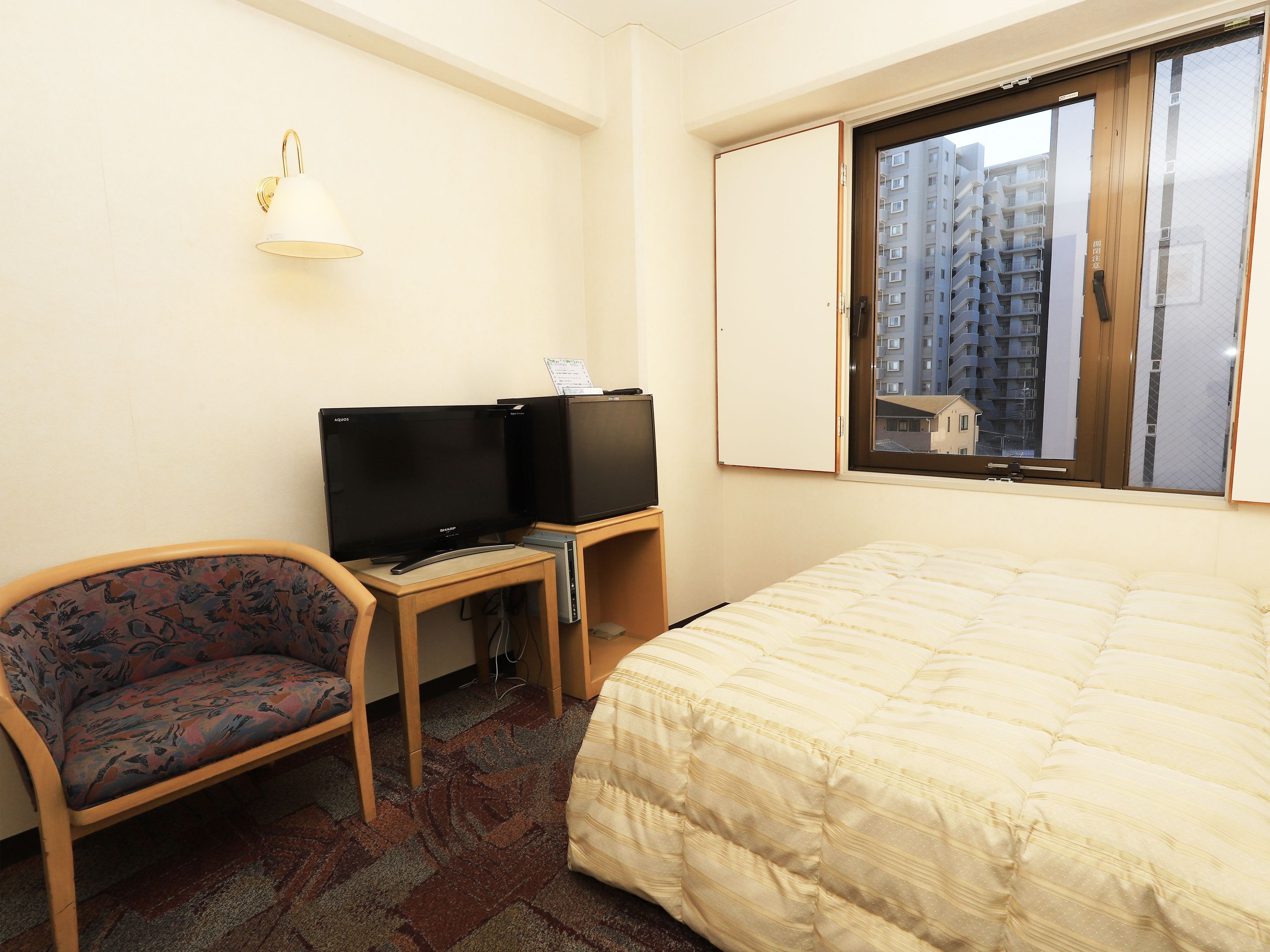 A hotel room with a bed, a desk with a computer and a TV, a chair, and a window with a view of buildings.
