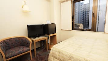 A hotel room with a bed, a desk with a computer and a TV, a chair, and a window with a view of buildings.