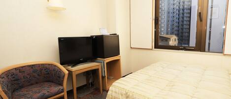 Habitación de hotel con cama, escritorio con computadora y televisión, una silla y ventana con vista a edificios.