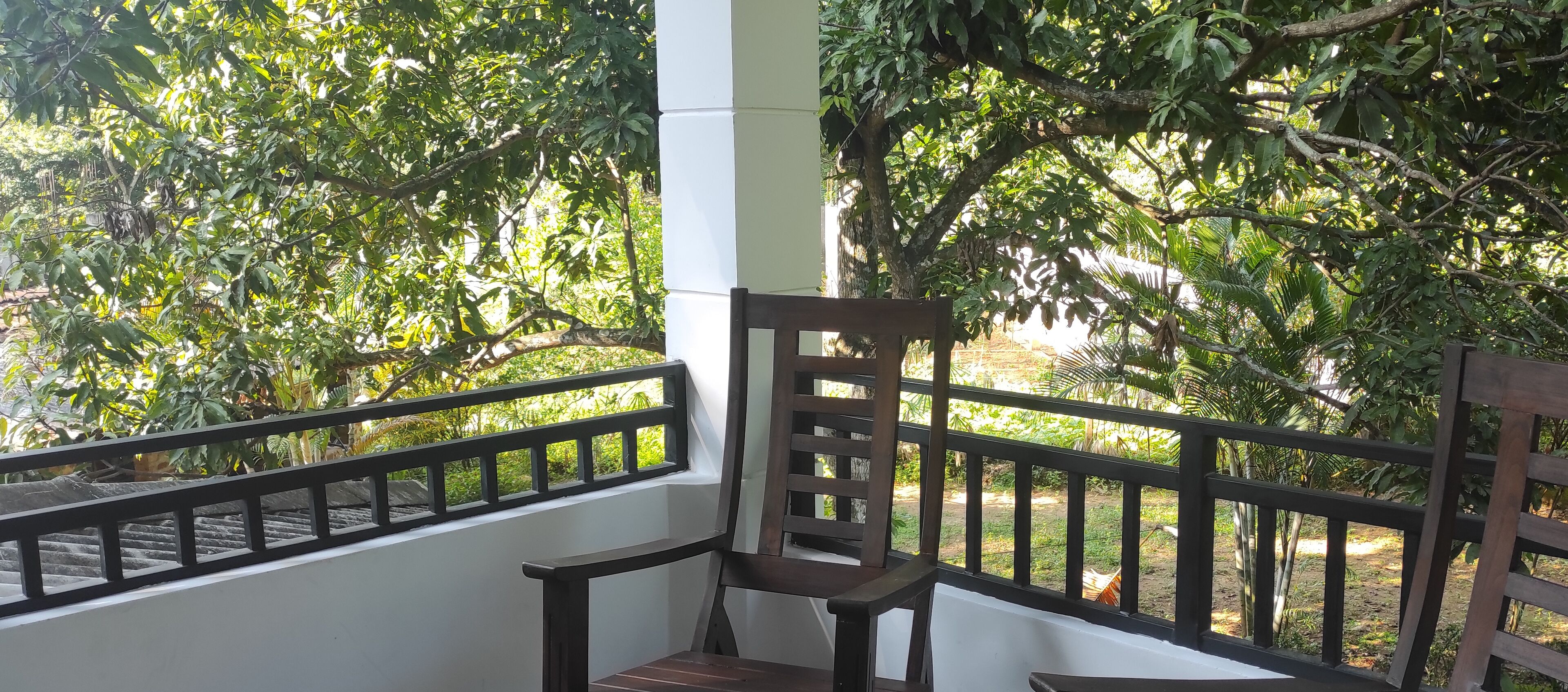 A wooden chair on a balcony with a black railing, overlooking a garden with various trees and plants.