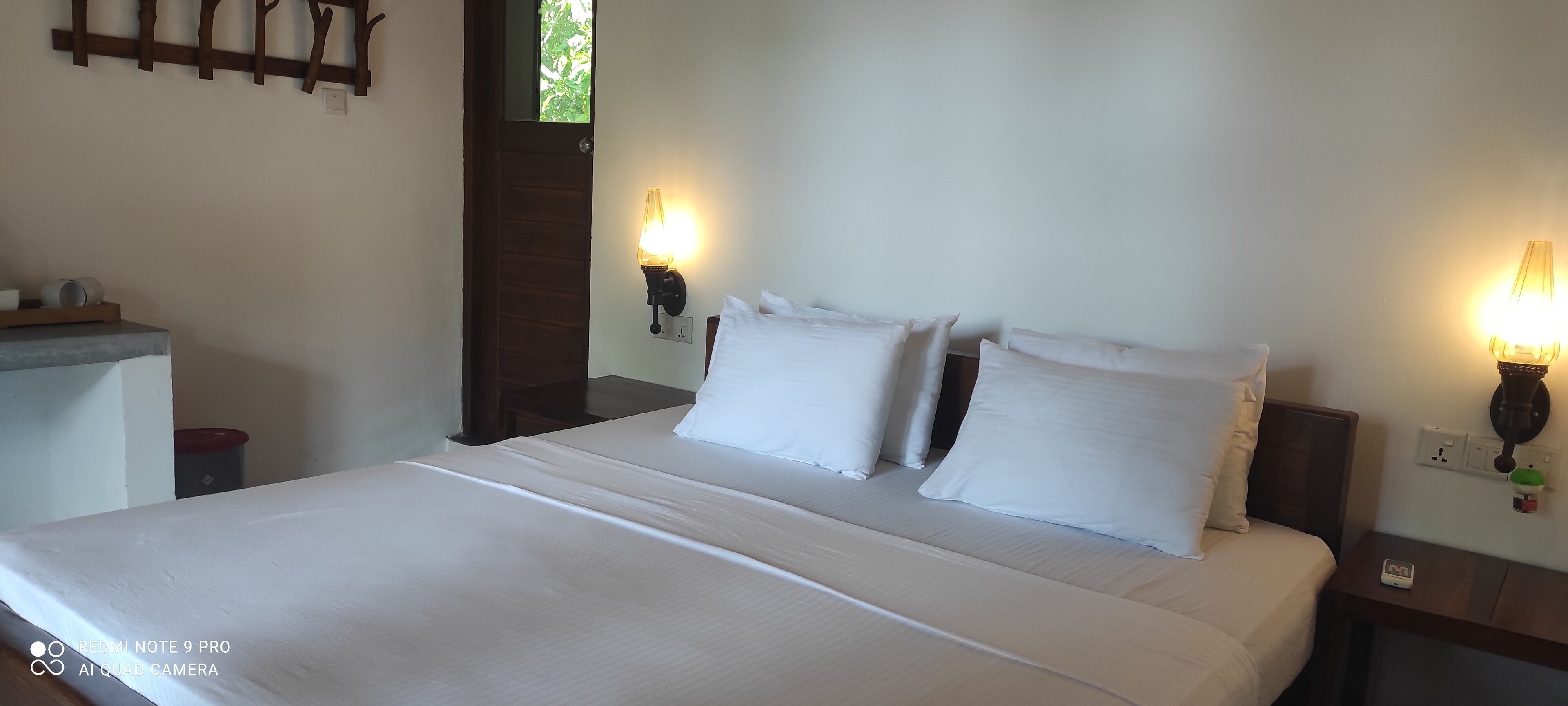 A neatly made bed with white linens and pillows, a bedside table with a lamp, and a wall-mounted light fixture.