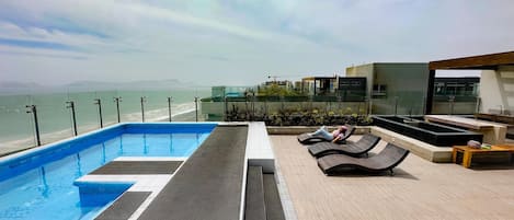 A rooftop pool area with a view of the sea, lounge chairs, and a modern architectural design.