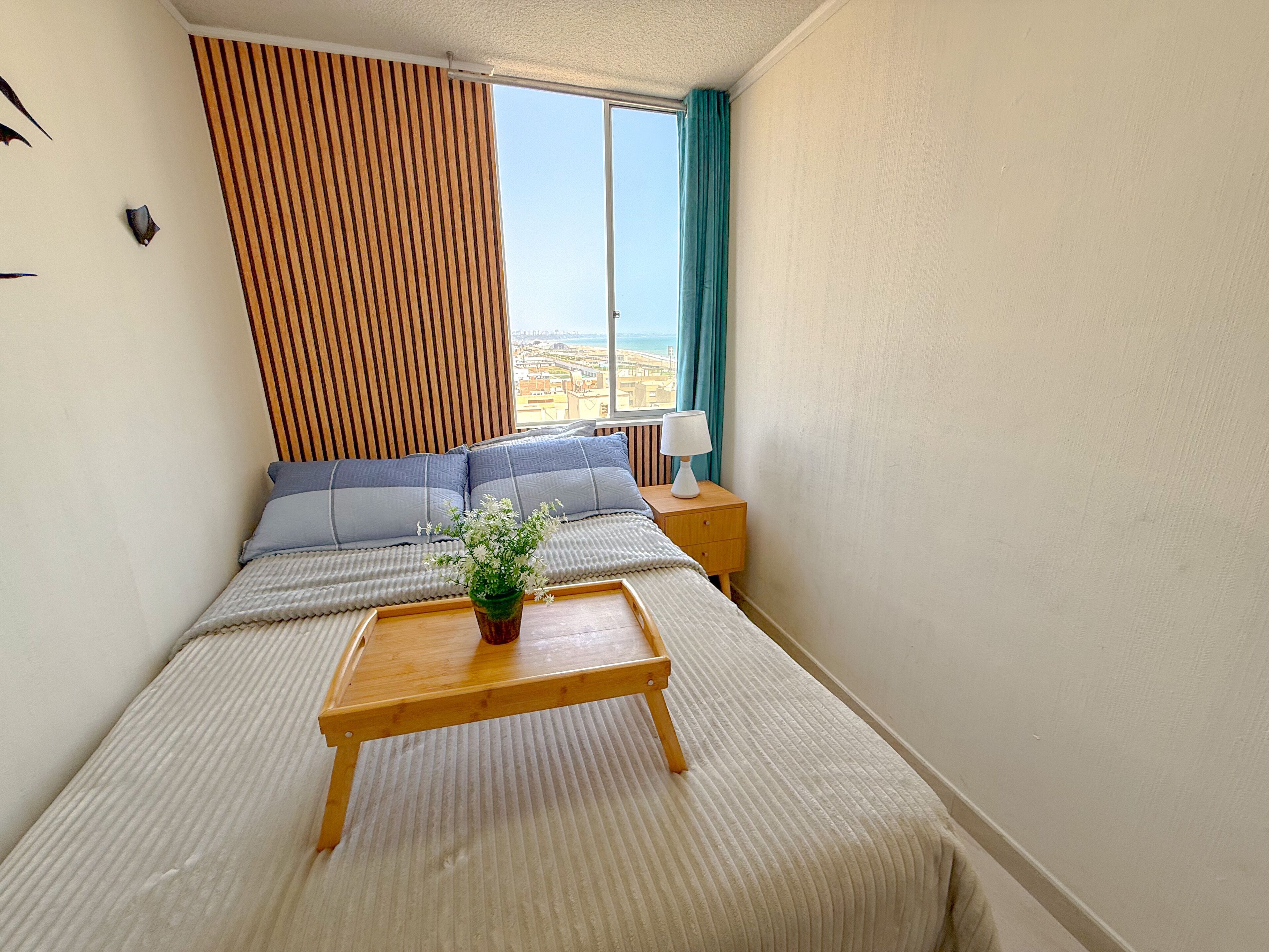Een netjes opgemaakt slaapkamer met een bed, een nachtkastje met een lamp, een raam met gordijnen en uitzicht op de stad.